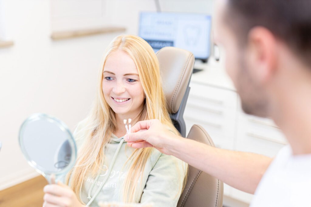 Junge blonde Frau bekommt das Nachher Ergebnis ihres Bleachings vom Zahnarzt gezeigt. Er hält die Vergleichsfarbenen Zähne vor ihr während sie sich im Spiegel betrachtet