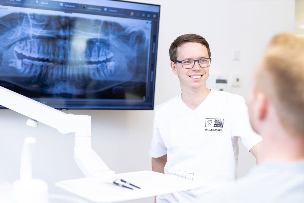 Zahnarzt bespricht mit einem Patienten, der auf dem Behandlungsstuhl sitzt das 3D Röntgenbild am Fernseher